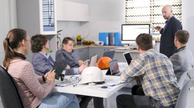 İnşaat şirketinin çalışanları konferans odasında bir masada oturuyorlar. Mimarlık Bürosu. Elinde kalem olan bir adam sunum yapıyor ve bir grup insan dikkatle dinliyor.