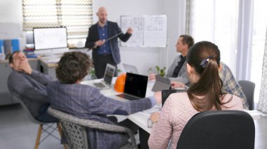 Bir grup iş adamı bir masada oturuyor ve takım elbiseli bir adam bir sunum yapıyor. İnşaat mühendisi elinde bir işaret tutuyor ve şirket personeli onu dikkatle dinliyor.