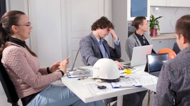 Mimarlık bürosunun yöneticileri yeni bir tanıtım kampanyası geliştiriyorlar. Bir grup insan toplantı odasında bir masada oturuyorlar. Yorgun bir adam başını tutar.