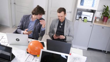 İki tasarım mühendisi toplantı odasında dizüstü bilgisayarlarıyla bir masada oturmuş cep telefonlarına bakıyorlar. Genç profesyoneller. Mimarlık bürosundaki brifing sırasında ara..