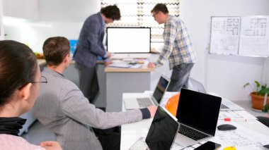 İki adam şirket personeline yeni bir mimari proje sunuyor. Bir grup insan bir konferans odasında birlikte çalışıyor. Masada birkaç dizüstü bilgisayar var. Atmosfer odaklı.
