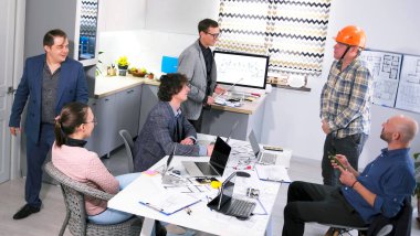 İnşaat şirketi çalışanları turuncu kasklı bir işçiyi dinliyor. Bir grup insan, iş merkezinin inşaat aşamalarını tartışmak için konferans odasındaki masanın etrafında toplandı..