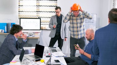 Şirketin bir çalışanı turuncu bir miğfer takar ve tesise girmeye hazırlanır. Bir grup adam dizüstü bilgisayarları, çizimleri ve planları olan bir masada oturuyor..