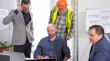 Bir grup inşaat mühendisi ortak bir proje üzerinde çalışıyor. Adamlardan biri yeşil bir güvenlik yeleği ve turuncu bir kask giyiyor. Laptop ekranına bakıyorlar..