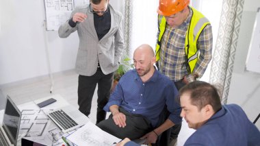 İnşaat müteahhiti proje tahminini şirketin yönetimiyle tartışıyor. Bir grup adam dizüstü bilgisayar ekranında bir binanın planlarına bakıyor. Mimarlık Bürosu