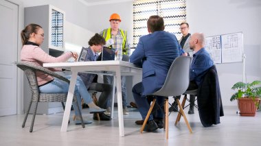 Yönetim kurulu proje tahminini müteahhitle tartışıyor. Bir grup iş adamı konferans odasında bir masada oturuyor. Atmosfer profesyonel ve odaklı.