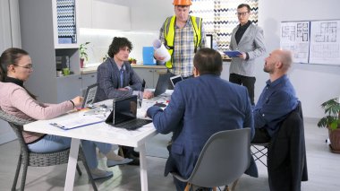 Yeşil yelekli bir inşaat işçisi ve turuncu bir şapka şirketin yönetimine yapılan işleri bildiriyor. Bir grup mimar ve mühendis konferans odasında bir masada oturuyorlar..