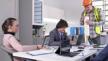 Bir grup inşaat alanı müşterisi proje tahminlerini ustabaşıyla koordine ediyor. Turuncu şapkalı ve yeşil yelekli bir erkek işçi elinde binanın çizimlerini tutuyor..