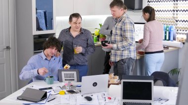 Meslektaşlar yemek yemek için rahat ofis mutfağındaki masanın etrafında toplandılar. Bazıları salata yiyor, sandviç yiyor ya da kahve içiyor. Atmosfer rahat, insanlar birbirlerinin arkadaşlığından zevk alıyor..