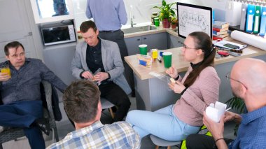 İş arkadaşları ofis mutfağındaki bir masada oturuyorlar, bazıları portakal suyu içiyor, sandviç yiyor, salata yiyor ve kahve içiyor. Atmosfer sakin, sohbet etmek için bir araya geldiler.