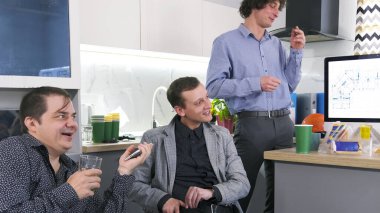 Bir grup adam arka planda bilgisayar monitörü olan bir ofis mutfağında bir masada oturuyorlar. Gülüyorlar ve birbirlerinin arkadaşlığından zevk alıyorlar. İş yemeği. Kahve molası.