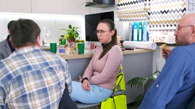 Genç bir kadın ofis mutfağında erkek meslektaşlarıyla sohbet ediyor. Kahve molası. İş yemeği. Bir grup insan ofis mutfağında bir masada oturuyor. Yanında oturan adam bir sandviç yiyor.