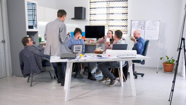 Bir grup başarılı profesyonel insan konferans odasında bir masada oturup yemek yiyorlar. Yemek yiyip içiyorlar. Masada birkaç dizüstü bilgisayar ve inşaat çizimleri var..