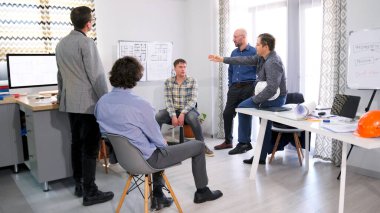 Bir grup adam modern bir iş merkezinin inşaat aşamalarını koordine ediyor. Mühendisler ve inşaatçılar proje tahminlerini tartışıyorlar. Sahne profesyonel ve odaklı.