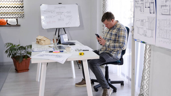 Дизайнер сидит за своим столом и болтает по мобильному телефону. Главный архитектор компании работает над новым строительным шедевром. У него есть чертежи на столе, ноутбуке и т.д..