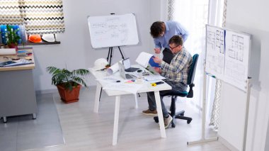 Mühendis proje tahminini muhasebeciyle tartışıyor. İki adam mimari çizimler üzerinde çalışıyor. Sahne, önemli bilgileri tartışan ya da plan yapan adamlarla odaklanmış ve ciddidir..