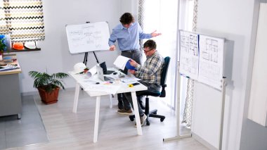Tasarımcı ve inşaat projesinin mühendisi çalışmanın ayrıntıları hakkında tartışıyorlar. İki adam, çizimler ve planlarla dolu beyaz bir masası olan mimari bir ofiste..