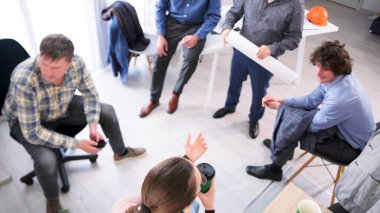 Çemberin içinde oturan bir grup insanın üst görüntüsü, içlerinden biri çizimlerle dolu bir rulo tutuyor. Sahne, iş arkadaşlarıyla birlikte bir proje üzerinde çalışırken ya da bir konuyu tartışırken işbirlikçi ve odaklıdır..