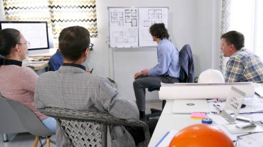 Bir grup insan, çeşitli binaların çizimleri ve planlarıyla bir sunum standının önünde oturuyor. Mühendisler, inşaatçılar ve tasarımcılar inşaat projesinin kârlılığını tartıştılar.