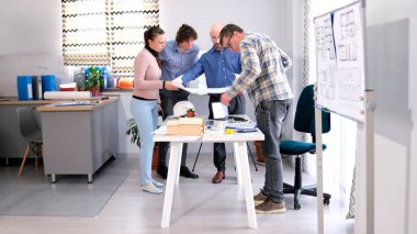 Bir grup inşaatçı bina çizimlerini inceliyor. Mimarlar konferans odasında bir masada toplandılar ve bir proje üzerinde çalışıyorlar. Sahne işbirlikçi ve odaklı.