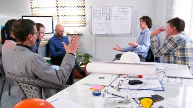 İnşaat şirketinin yöneticileri projenin kârlılığını tartıştılar. Bir grup insan, çizimler ve inşaat planlarıyla bir masa ve tezgah etrafında toplandı. Sahne ciddi ve odaklanmış.