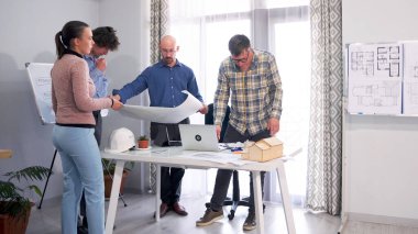 Bir grup mühendis, içinde çizimler, bir kır evi modeli ve bir dizüstü bilgisayar olan bir masanın etrafında toplandı. Mimarlar inşaat planlarını ve proje tahminlerini tartışıyorlar..