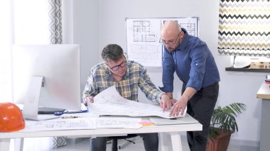 Mimarlık şirketinin müdürü inşaat projesi çizimlerinin detaylarını baş mühendis ile koordine ediyor. Gözlüklü erkek meslektaşlar ortak bir proje üzerinde çalışıyorlar.