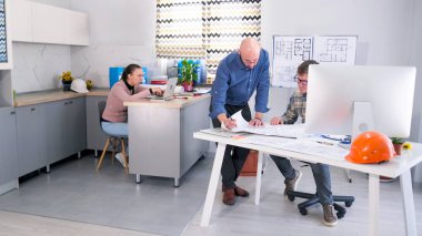 Bir adam, çizimleri olan bir mimarın masasında duruyor. Baş mühendis ve tasarımcı projenin maliyet tahminleri ve ayrıntılarını tartıştılar. Arkalarında bir kadın sekreter oturuyor..