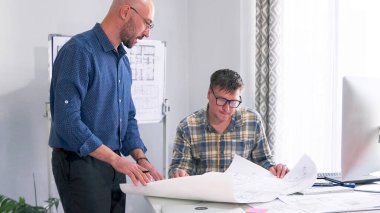 Mimar ve tasarımcı ortak bir projede birlikte çalışıyorlar. İki adam, iş merkezinin tahmini detaylarını ve inşa aşamalarını koordine ediyor. İş arkadaşları iş başında.