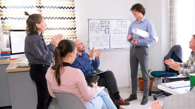 Bir grup insan başarılı bir genç öğretim üyesini alkışlıyor. Erkek bir mimar yeni inşaat şaheserinin sunumunu yapıyor. Sahne işbirliği ve takım çalışmasını temsil ediyor