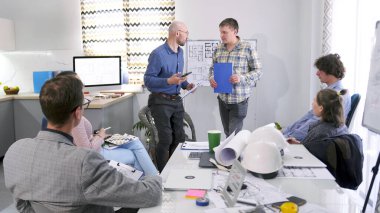 İki erkek mühendis bir sunum yapıyor. Bir grup insan konferans odasında bir inşaat projesini tartışıyor. Sahne profesyonel ve odaklı.