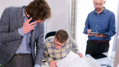 Üç adam mimari bir nesne için planlar üzerinde çalışıyor. Biri telefonda konuşuyor, diğeri de tabletten bir şey izliyor. Sahne odaklı ve ciddi.