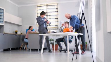 Şirketin baş mühendisi proje detaylarını müşterilerle koordine ediyor. Bir grup erkek, bir mimarlık firmasının aile ofisinde birlikte çalışıyor. Arka planda bir sekreter oturuyor..