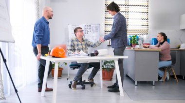 Baş mimar müşteriyi karşılıyor. Proje detaylarının koordinasyonu. Genç bir iş adamı gözlüklü bir adamla tokalaşıyor. İnşaat şirketinin ofisi.