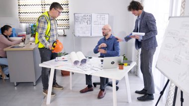 İnşaat şirketinin yöneticisi ve ustabaşı baş mimarla iletişim kuruyor. Bir grup adam bir proje üzerinde çalışıyor. Arkalarında bir kadın sekreter oturuyor..