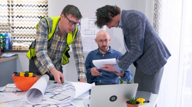 Üç odaklı adam, modern bir iş merkezi projesinin çizimleri, planları ve belgeleriyle çalışıyor. Mimarlar proje tahminlerini tartışıyor ve her şeyin yolunda olduğundan emin oluyorlar