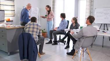 Bir adam ve bayan asistanı bir sunum yapıyorlar. Bir grup insan, arka planda çizimler ve planlarla dolu beyaz bir panoyla konferans odasında toplandı. Ekip projeyi tartışıyor.