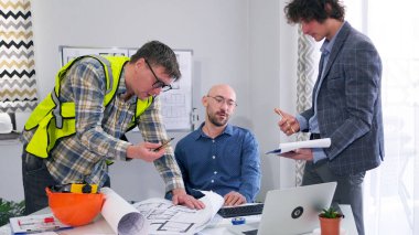 Mimarlar, inşaatçılar ve mühendislerden oluşan profesyonel bir ekip modern bir binanın çizimleri ve planları üzerinde çalışıyor. Üç adam inşaatın ayrıntılarını ve aşamalarını tartışıyor..