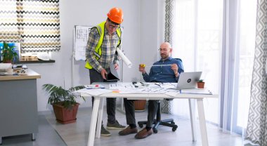 İnşaat şirketinin ofisinde iki adam var. Biri turuncu kask ve yeşil yelek giyiyor. Ustabaşı ve mühendis mimari nesnenin çizimlerini ve planlarını tartışıyor.