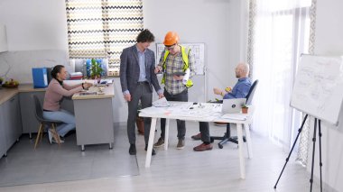 Yeşil yelek ve turuncu kasklı bir ustabaşı ve bir mühendis projenin detaylarını koordine etmek için patrona geldi. Bir grup insan çizimleri ve bina planlarını tartışıyor..