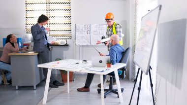 Turuncu şapkalı ve yeşil yelekli bir ustabaşı elinde planlarla konferans odasındaki patrona rapor veriyor. Arkalarında bir tasarımcı duruyor, kahve içiyor ve bir bayan sekreter oturuyor..