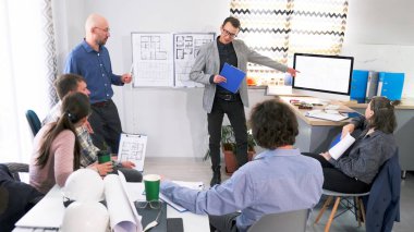 Profesyonel bir yönetici, bir grup insanın önünde mimarlık ve inşaat üzerine bir sunum yönetiyor. Öğretim görevlisi binanın çizimi olan bir bilgisayar monitörünü işaret ediyor. Sahne profesyonel ve odaklı.