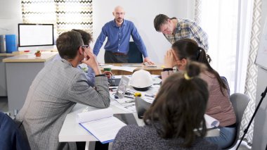 Mavi gömlekli ve gözlüklü bir adam bir grup çalışanın başında duruyor ve çalışanlardan birini azarlıyor. Brifing. Takım çalışması. Bir grup insan konferans odasında bir masada oturuyor..