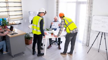 Bir grup inşaatçı projenin detaylarını mühendis ile tartışıyor. Yeşil yelekli iki adam elinde planlarla bir masanın etrafında duruyorlar. Şirket personeli planları tartışıyor.