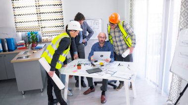 Mimarlık firmasının ekibi baş mühendisin elindeki laptop ekranına bakıyor. Bir grup inşaat işçisi ve planlamacı bir masanın etrafında toplanarak projenin planlarını tartıştılar..