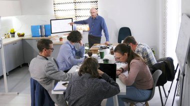 Bir grup insan konferans odasında bir masada oturuyor ve bir sunum izliyor. Adam ekranı işaret ediyor. Toplantı ya da brifing. Sahne odaklı ve ciddi, grup bir proje üzerinde çalışıyor.