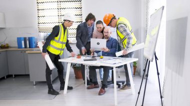 İnşaat şirketinden meslektaşlar dizüstü bilgisayara dikkatle bakıyorlar. Proje detaylarını tartışacağız. Bir grup inşaat işçisi ve mühendis, çizimler ve dizüstü bilgisayarla bir masanın etrafında toplandı.