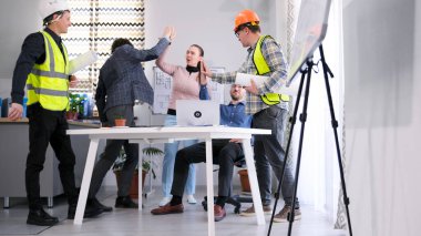 Bir grup inşaatçı, bir mühendis, bir mimar ve bir tasarımcı iyi bir işi kutluyorlar. Meslektaşlar beşlik çakıyor. Masanın etrafında dizüstü bilgisayar ve çizimlerle duruyorlar.