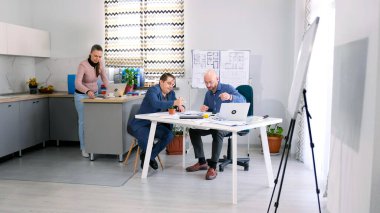 Üç ofis çalışanı bir inşaat projesi üzerinde çalışıyor. İki adam projenin planlarını, planlarını ve bütçesini tartıştı. Sahne işbirlikçi ve odaklı. Mimarlık Bürosu