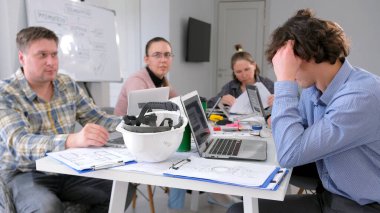 Bir grup ofis çalışanı konferans odasındaki bir masada, arkasında diyagramı olan beyaz bir pano ile oturuyorlar. Takım çalışması, takım oluşturma, beyin fırtınası. Toplantı